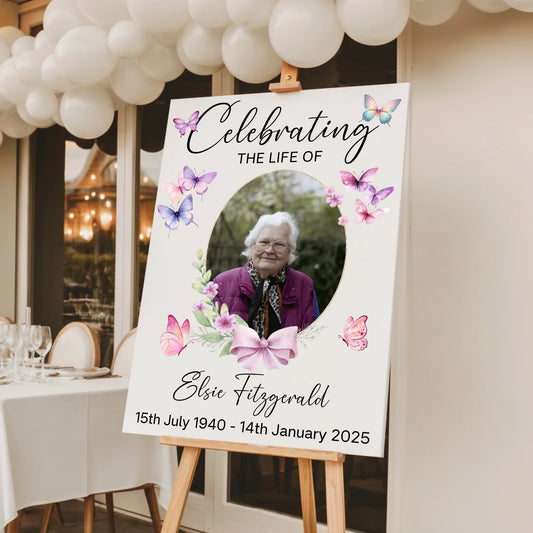 Personalised celebration of life memorial sign featuring a floral butterfly design with a photo of a loved one, custom name and dates, printed on white foam board and displayed on a wooden easel for a funeral or memorial service.
